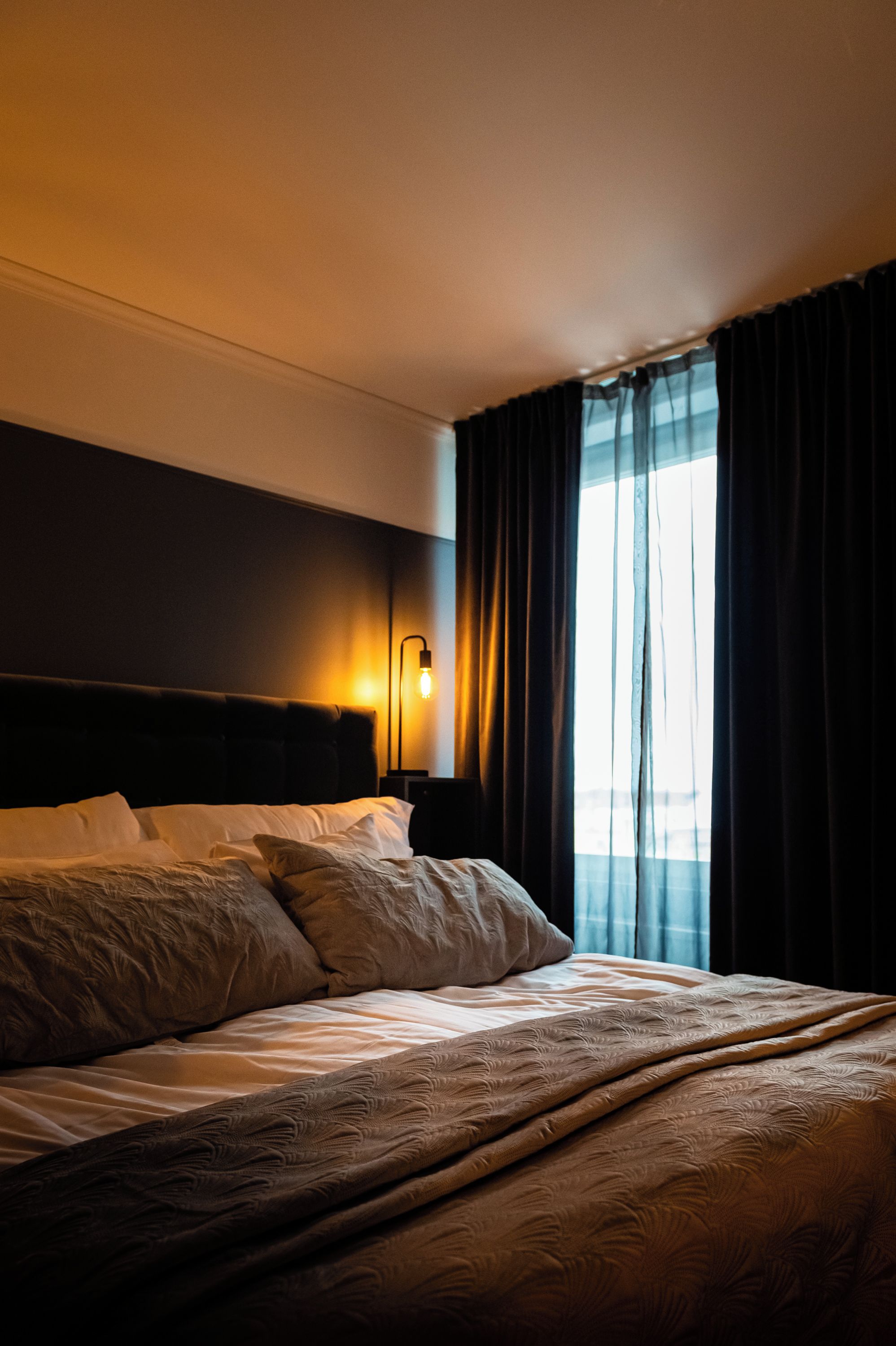 Ein Hotelzimmer mit einem großen Bett, einem Nachttisch, einer Wandlampe und einem Fenster mit Vorhängen.