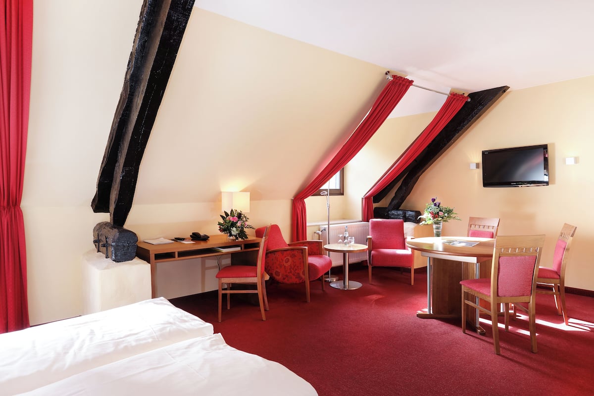 Habitación de hotel con cama, escritorio, silla, mesa, televisor y ventana con cortinas.