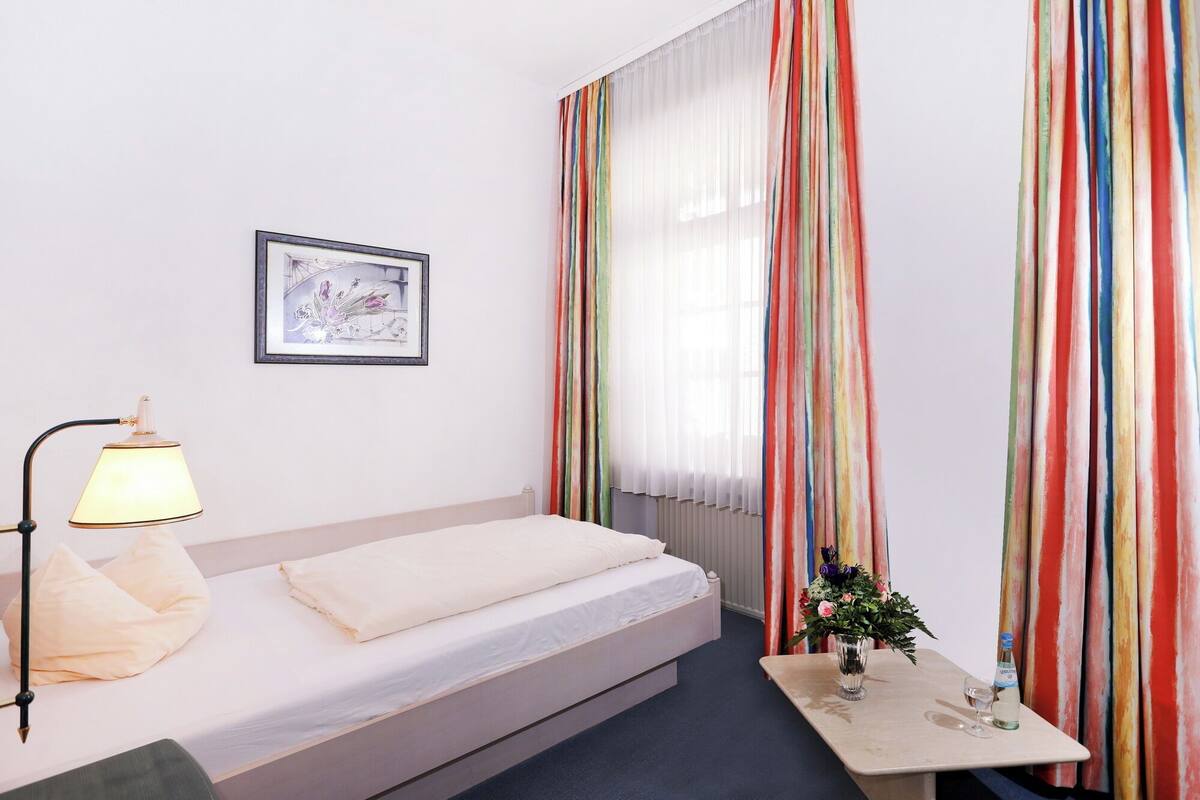 Una cama bien hecha con edredón blanco y almohadas, una lámpara de noche, un cuadro en la pared y una mesita con un jarrón y una botella.