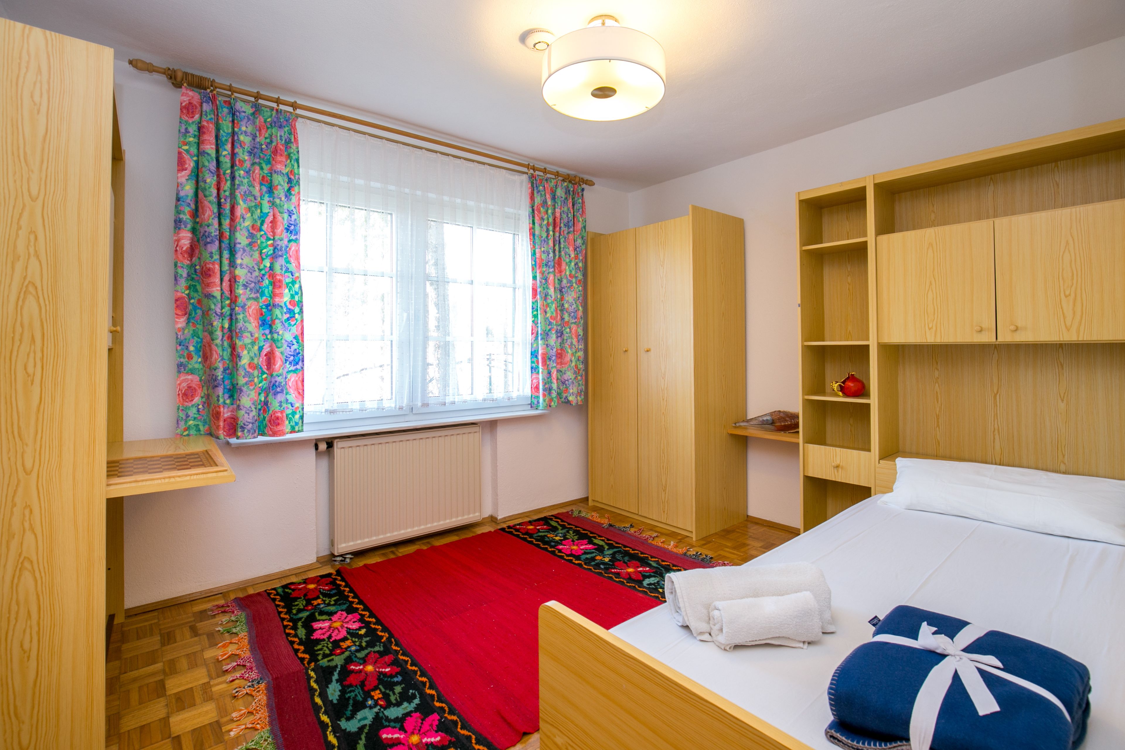 Un dormitorio con una cama de madera, un tapete floral en rojo y negro, un armario de madera y una ventana con cortinas florales.