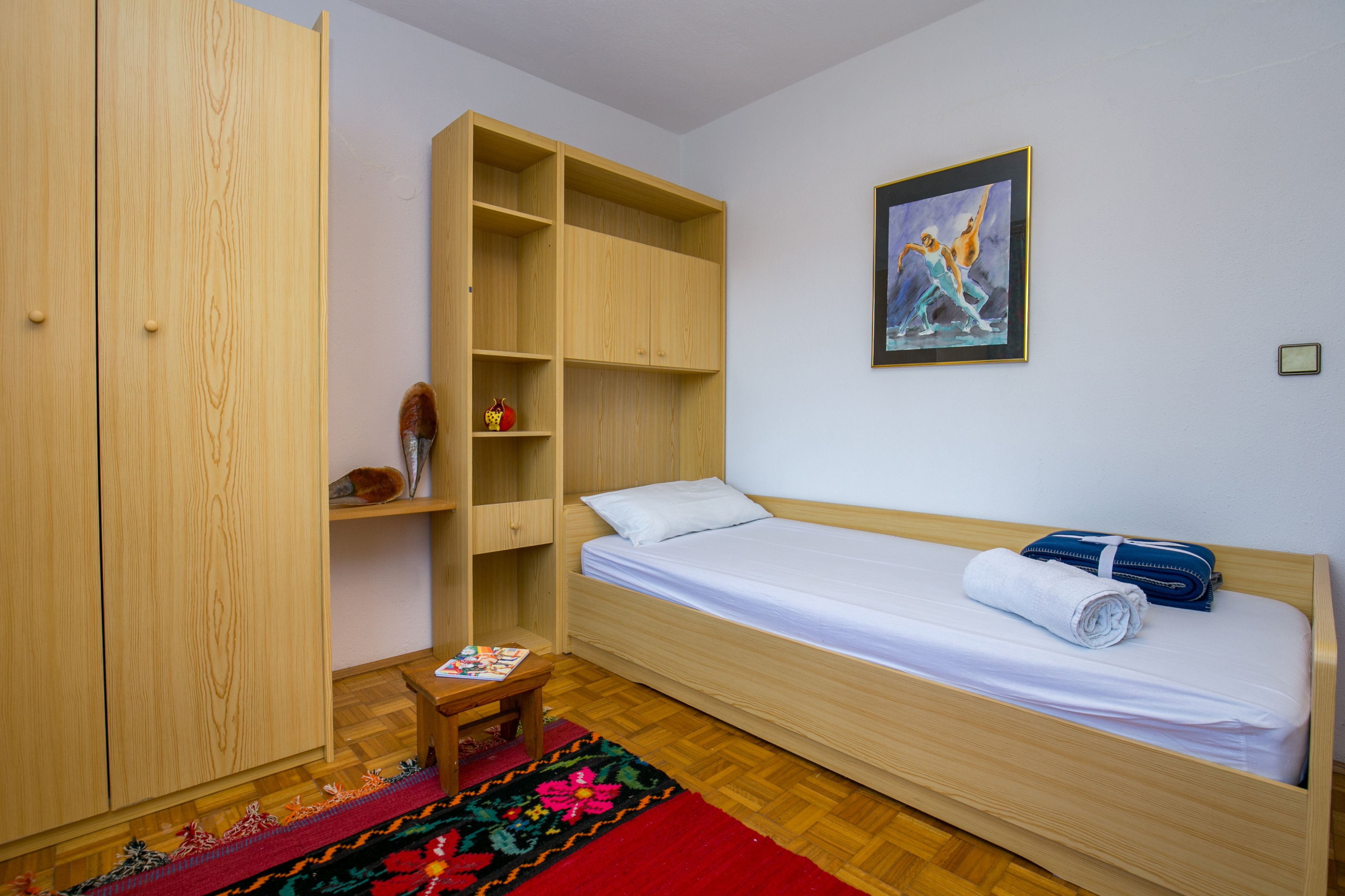 A room with a wooden wardrobe, a bed with a blue bag, a painting on the wall, and a colorful rug.