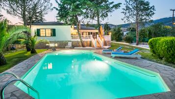 A swimming pool with a stone deck, a house with a balcony, and a lawn with a chair and a sun lounger.