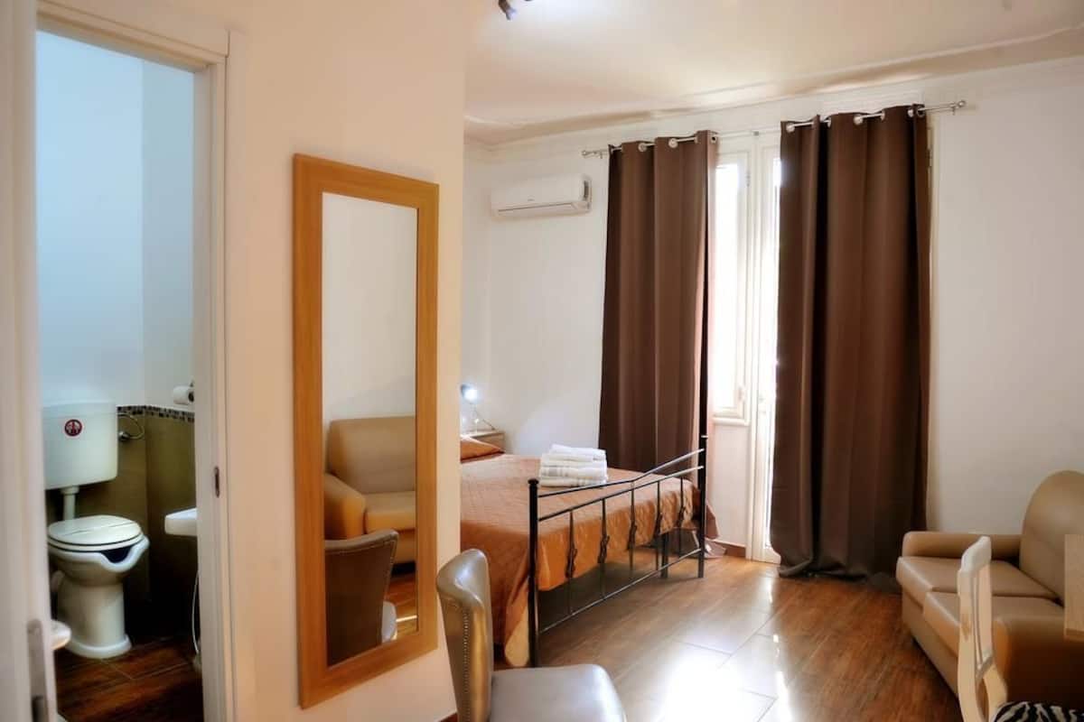 A bedroom with a bed, a chair, a mirror, a window with curtains, and a bathroom with a toilet and sink.