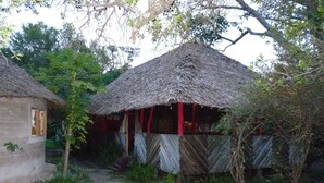 A rustic thatched-roof structure with a wooden door and a small window, surrounded by greenery and a dirt path.
