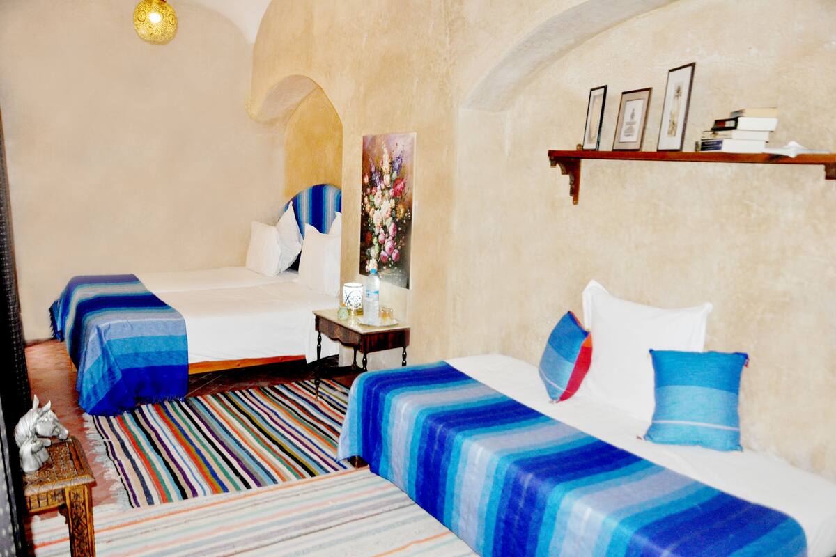A hotel room with two beds, a striped rug, a small table with a lamp, and a shelf with framed pictures.