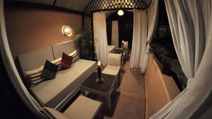 A cozy outdoor seating area with a cushioned bench, a small table with a candle, and a decorative wall light. - Riad Hidden (Marrakech)