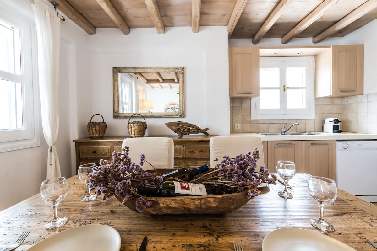 Une cuisine rustique avec des meubles de rangement en bois, une table à manger en bois sur laquelle se trouve un saladier rempli de vin et des verres, et un miroir accroché au mur.