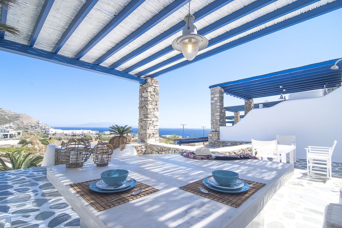 Un espace extérieur lumineux, aménagé pour les repas, avec des tables en pierre et de la vaisselle bleue, offrant une vue sur un paysage côtier.