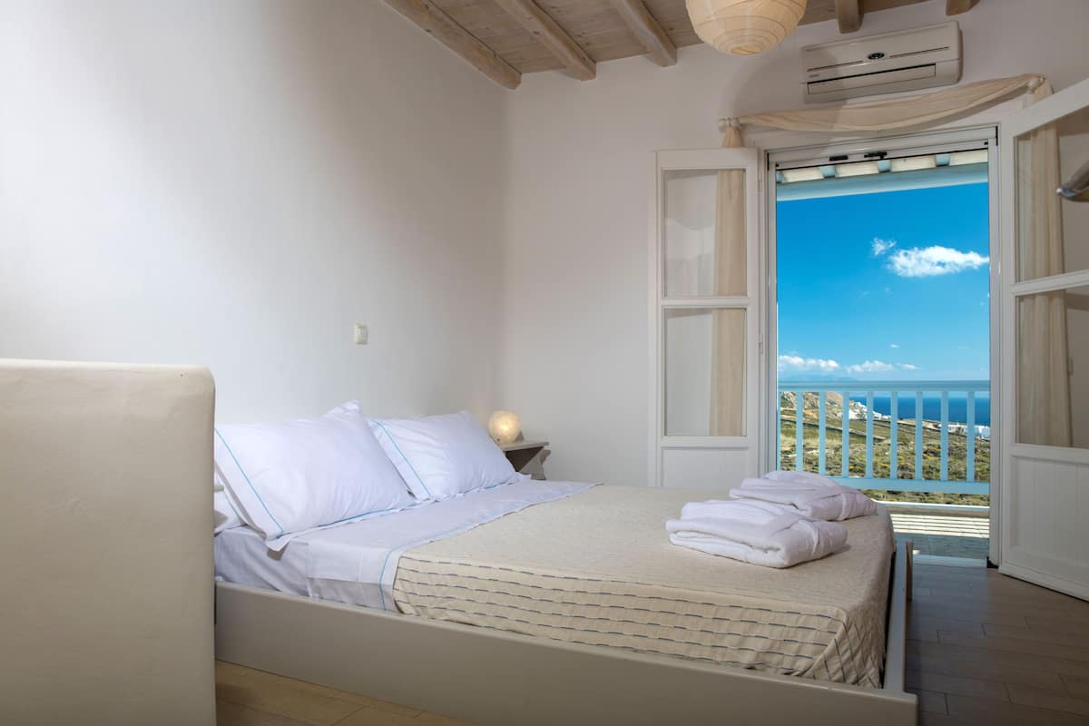 Une chambre à coucher avec un lit, une table de chevet et une vue sur la mer, visible à travers une porte ouverte.