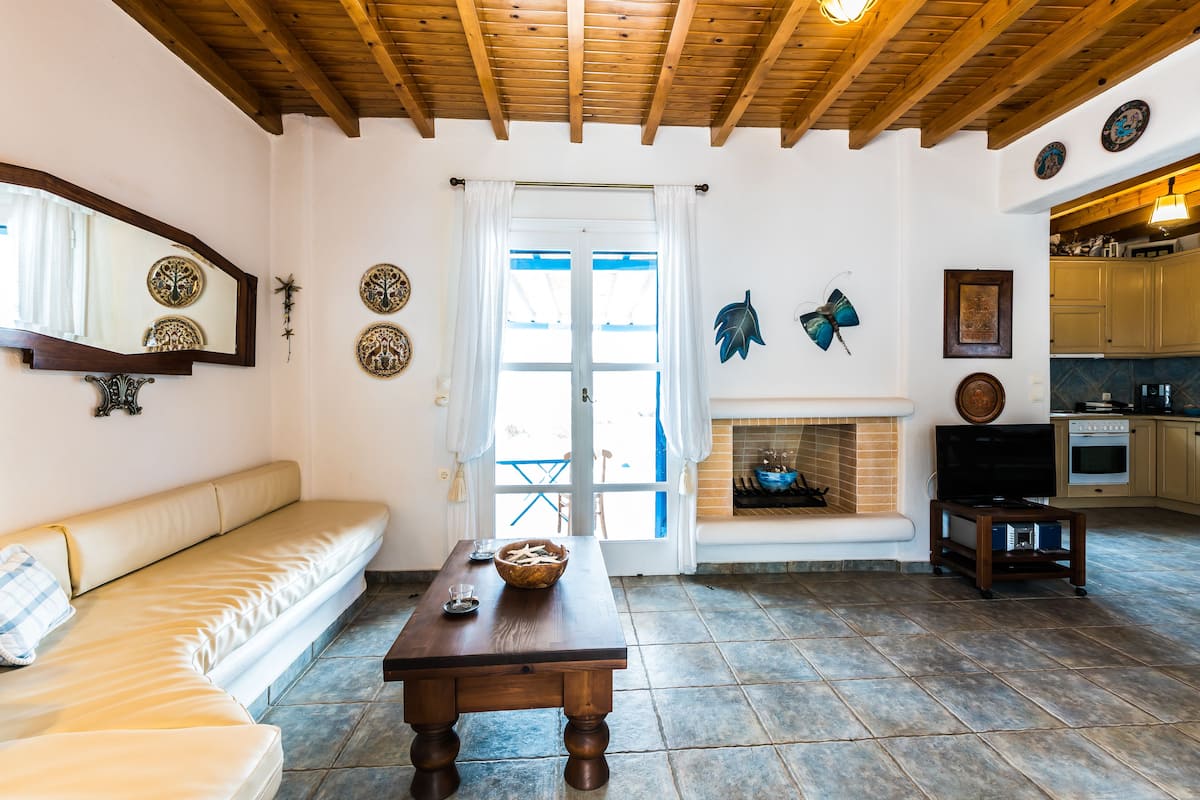 Un salon avec un plafond en bois, un canapé, une table avec un saladier, une cheminée et un coin cuisine.