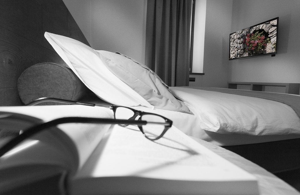 A bedroom with a bed, pillows, a bedside table with glasses, a window with curtains, and a wall-mounted painting.