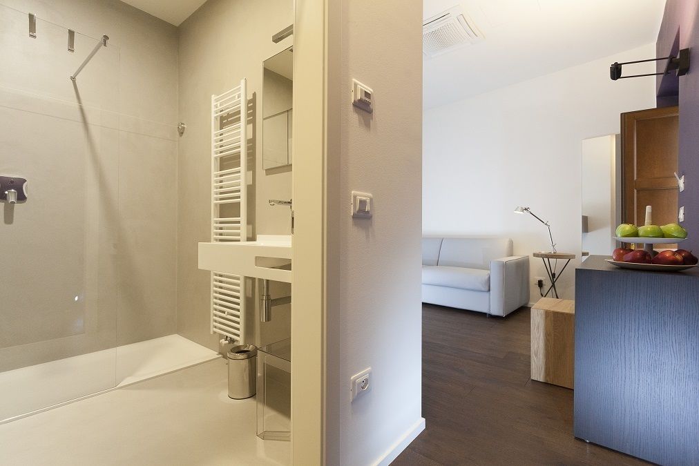 A modern bathroom with a shower, toilet, and towel rack. There is a white sink with a mirror above it and a small table with a lamp and fruit bowl. A white sofa is visible in the background.