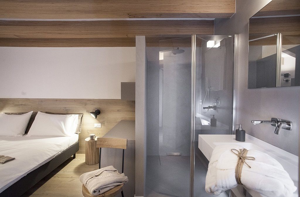 Una habitación de hotel moderna con cama, mesita de noche, lámpara y zona de ducha con paredes de vidrio.