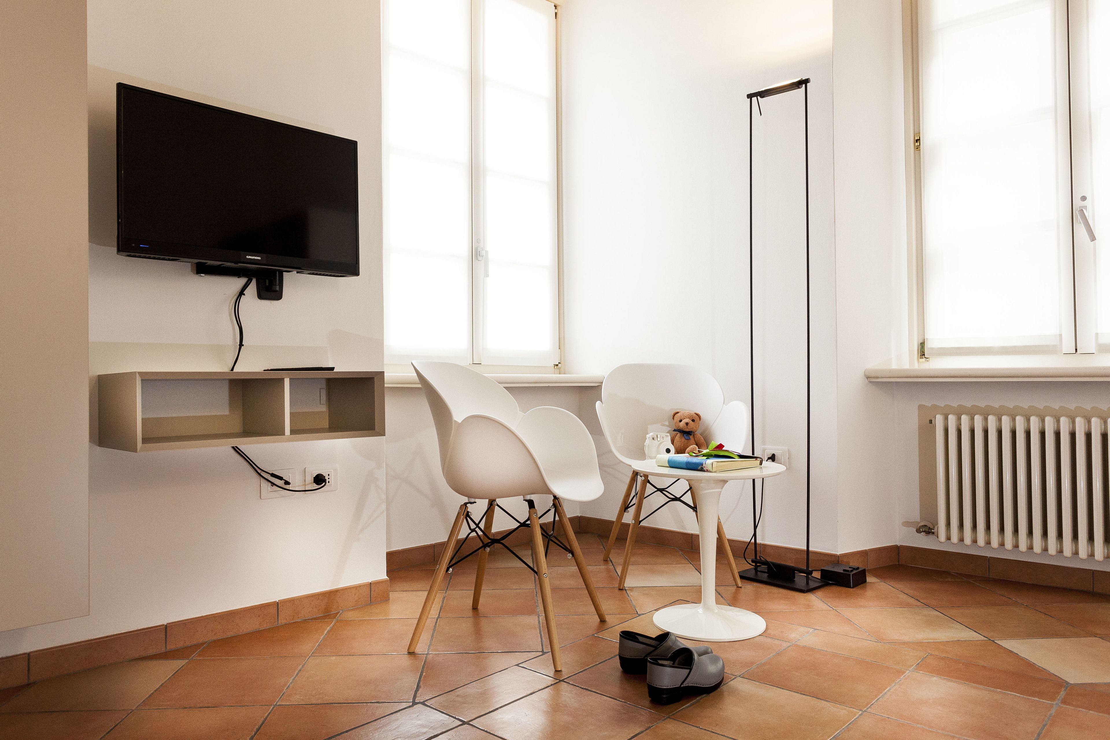 A modern room with a flat-screen TV mounted on the wall, a small table with a teddy bear, white chairs, and a floor lamp.