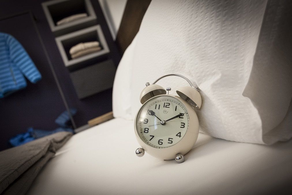 Un despertador blanco sobre una cama con un almohadón.