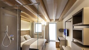 Una habitación de hotel moderna con cabina de ducha de vidrio, cama con cabecero de madera, televisor de pantalla plana montado en la pared y escritorio con silla.