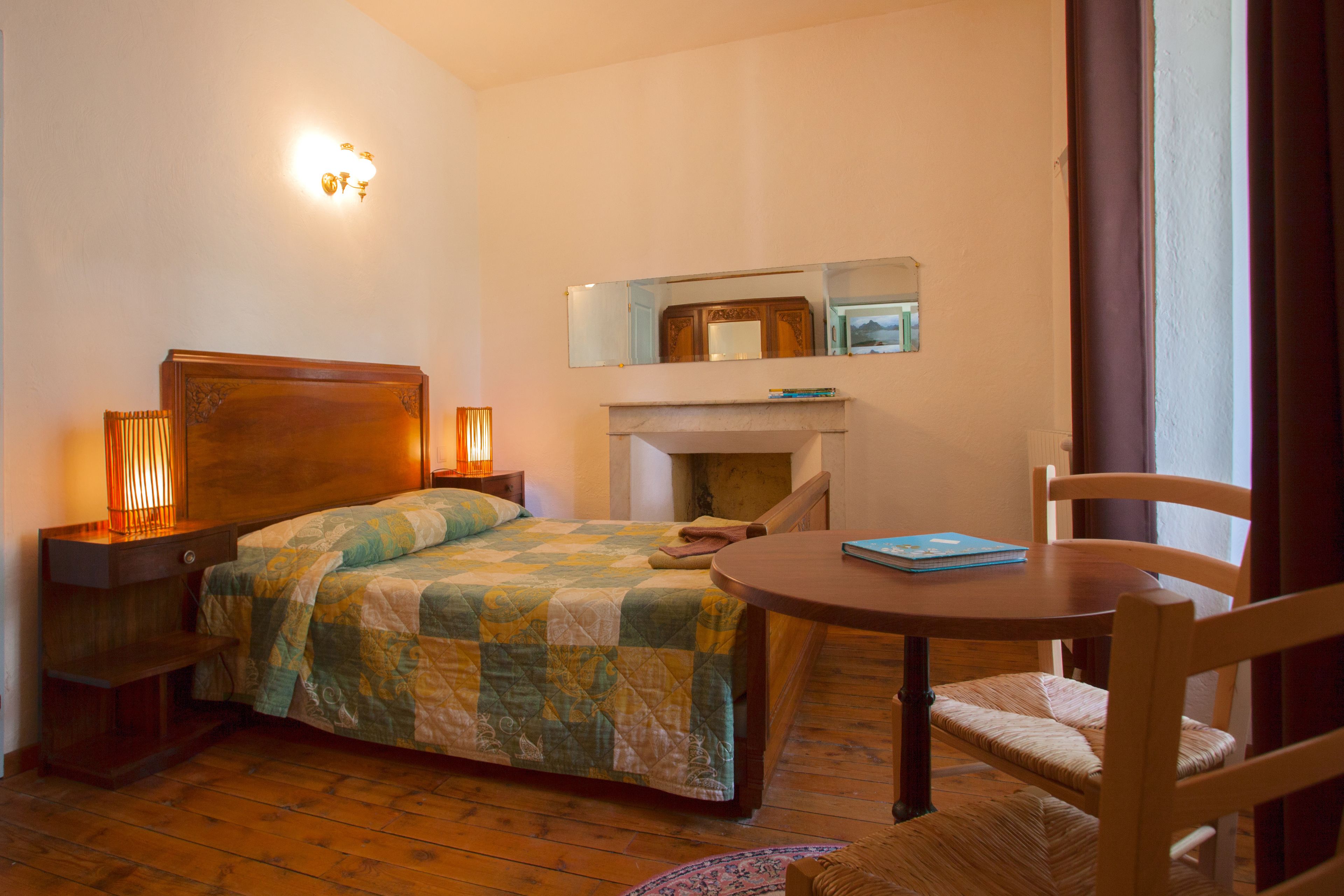 A bedroom with a bed, bedside lamps, a round wooden table, a chair, a mirror, and a fireplace.
