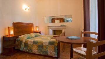 A bedroom with a bed, bedside lamps, a round wooden table, a chair, a mirror, and a fireplace.