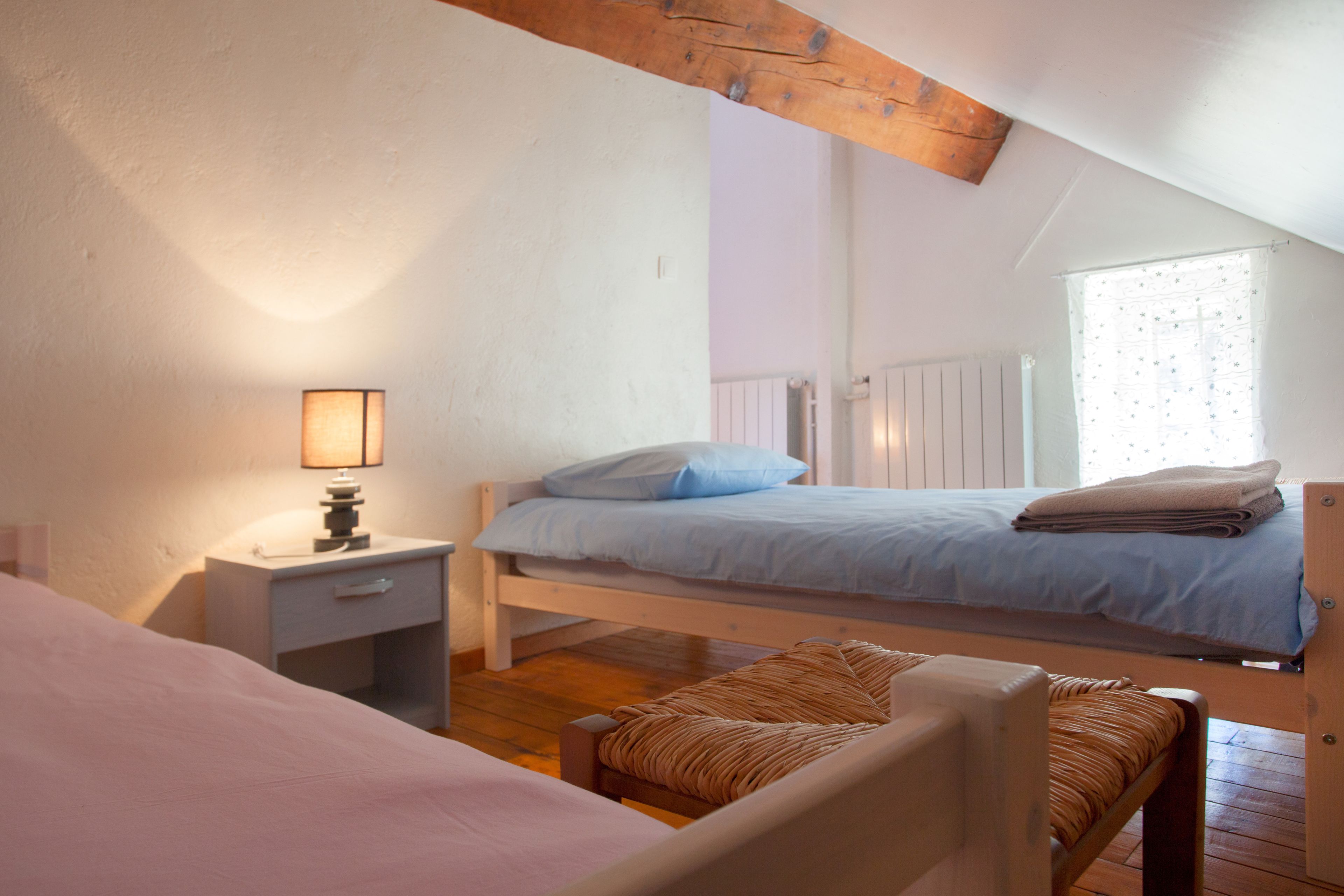 A bedroom with two beds, a nightstand, a lamp, a window with curtains, and wooden beams on the ceiling.