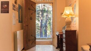 Un couloir avec une porte en bois donnant sur un espace extérieur arboré. Il y a un radiateur à gauche, une lampe à droite et un meuble avec des tiroirs.
