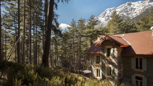 Une maison de campagne en pierre, avec un toit recouvert de tuiles rouges, entourée de grands pins et dominée, au loin, par une montagne enneigée.