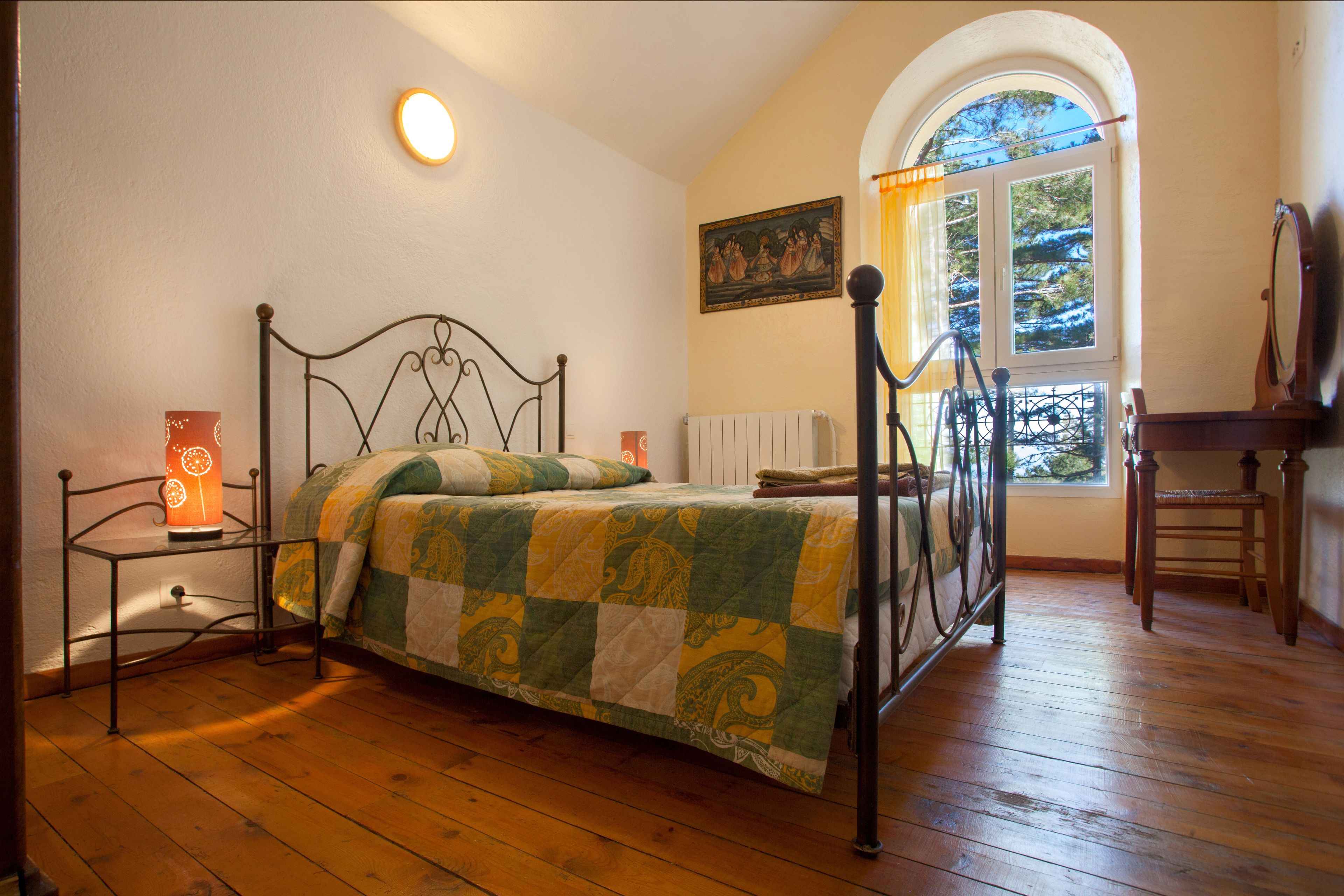 A bedroom with a bed, a bedside table with a lamp, a chair, and a window with a view of trees.