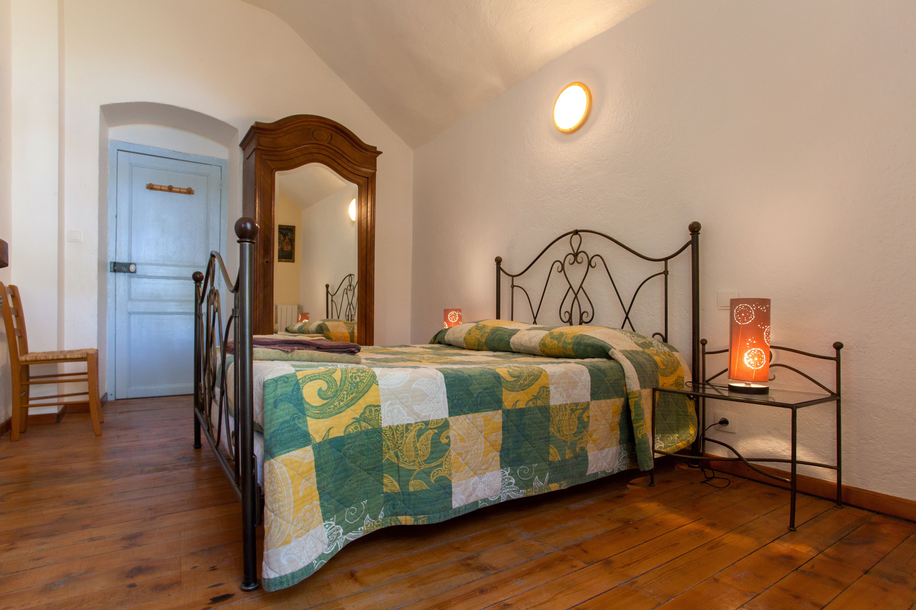 A bedroom with a bed, a chair, a bedside table with a lamp, a door, and a mirror.