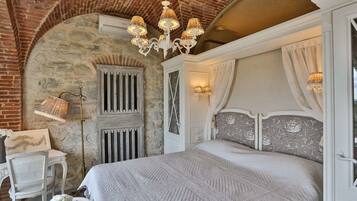Camera da letto con parete in pietra, un letto con testiera decorata, un lampadario e una finestra con tende.