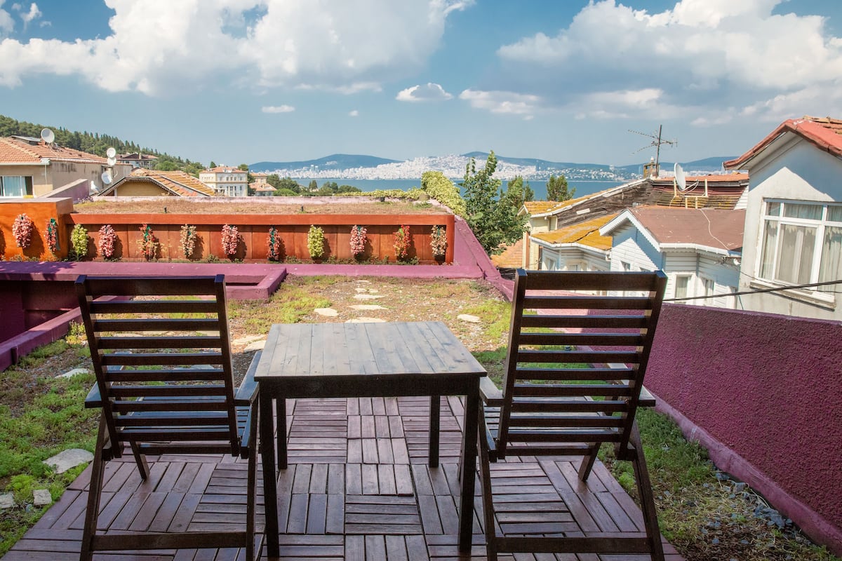 Una terraza de madera con dos sillas y una mesa, con vista a un paisaje urbano que incluye edificios y montañas.