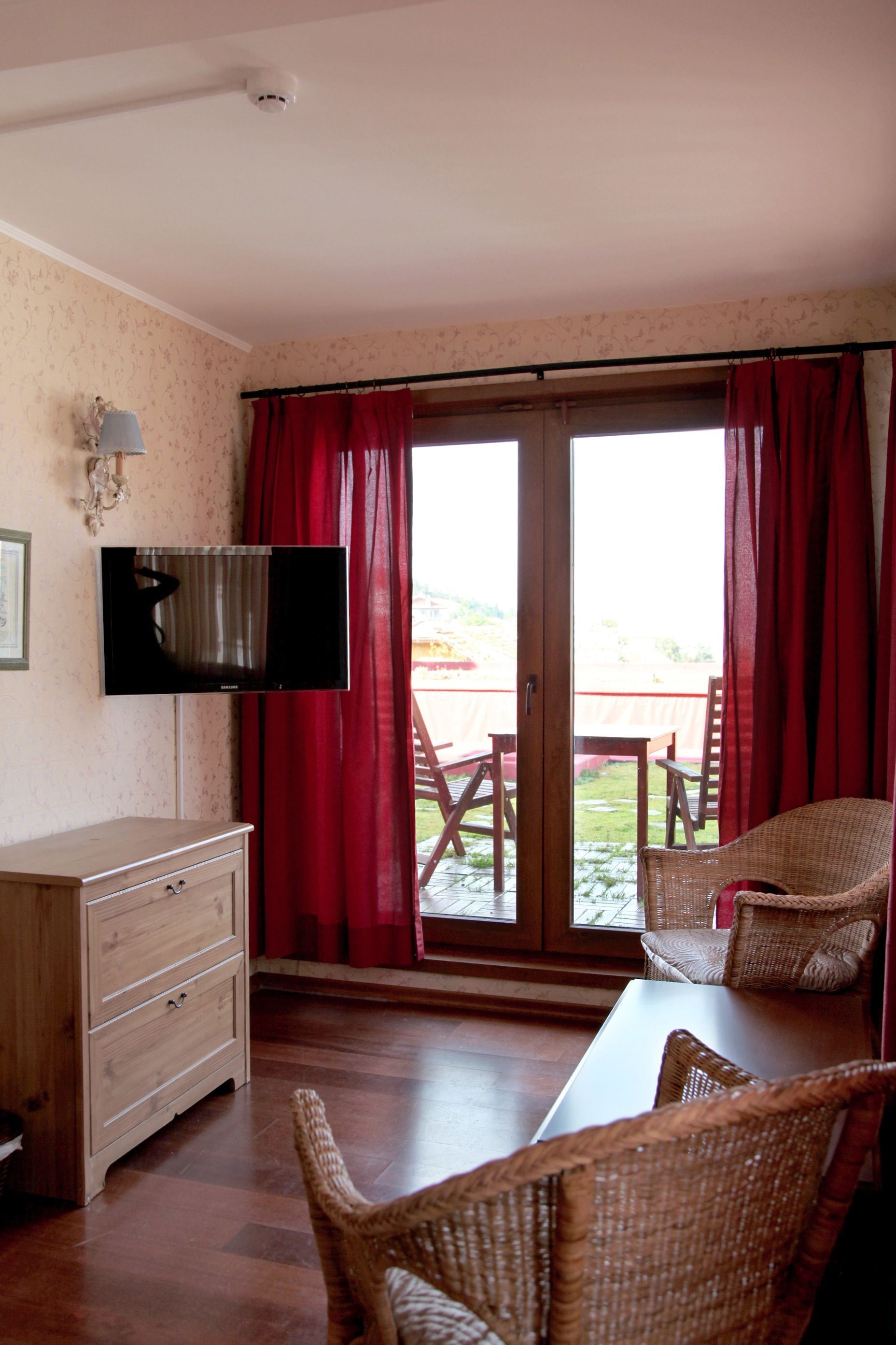 A room with a television mounted on the wall, a wooden dresser, wicker chairs, a table, and a sliding glass door leading to an outdoor area with a wooden table and chairs.
