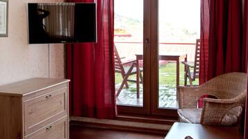 A room with a television mounted on the wall, a wooden dresser, wicker chairs, a table, and a sliding glass door leading to an outdoor area with a wooden table and chairs.