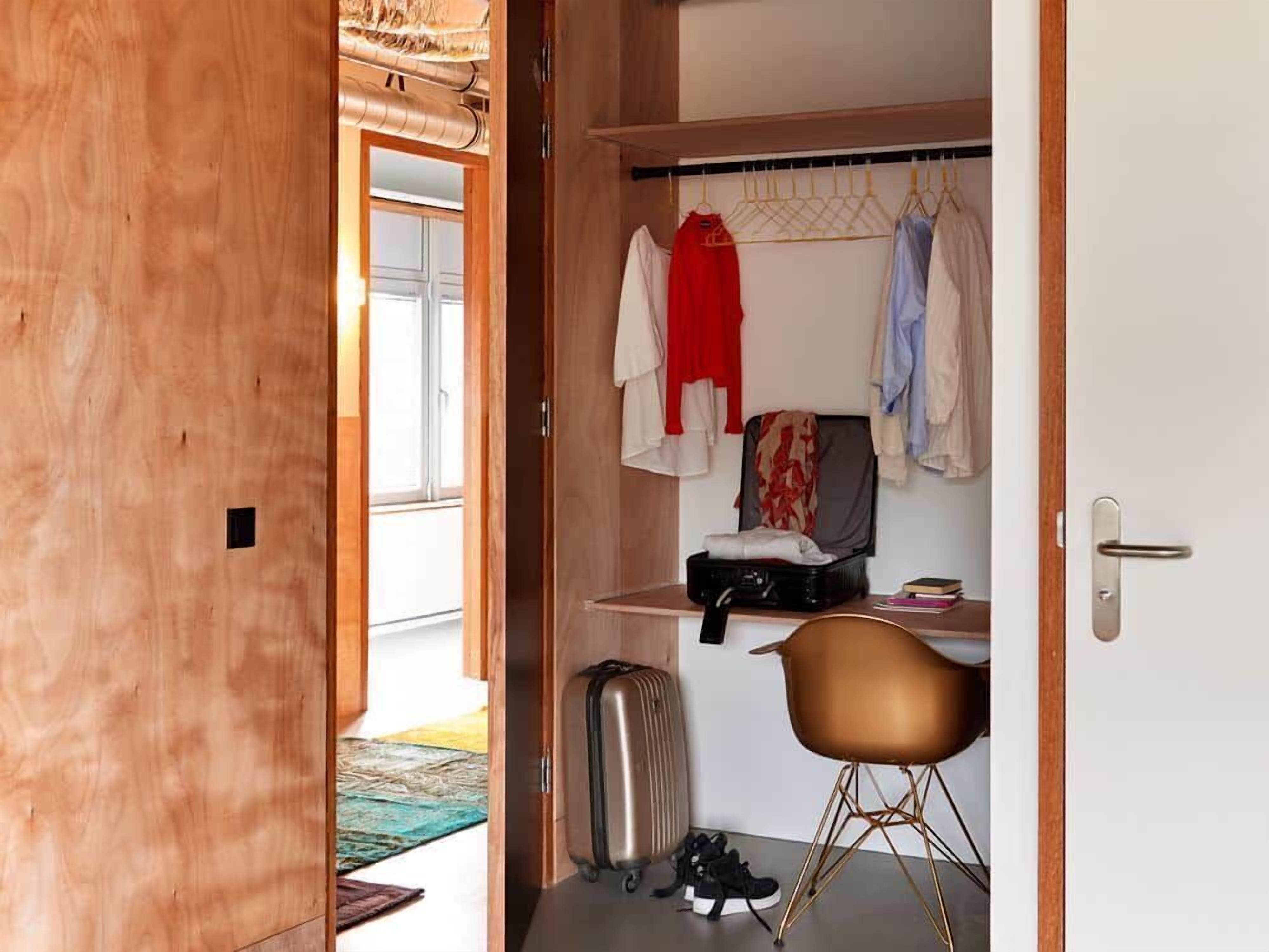 A compact room with a wooden wardrobe, a desk with a chair, a suitcase, and a hanging clothes rack.