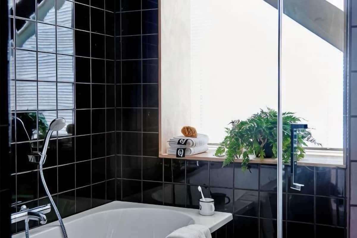 A modern bathroom with black tiles, a white bathtub, a glass shower enclosure, and a shelf with towels and a plant.