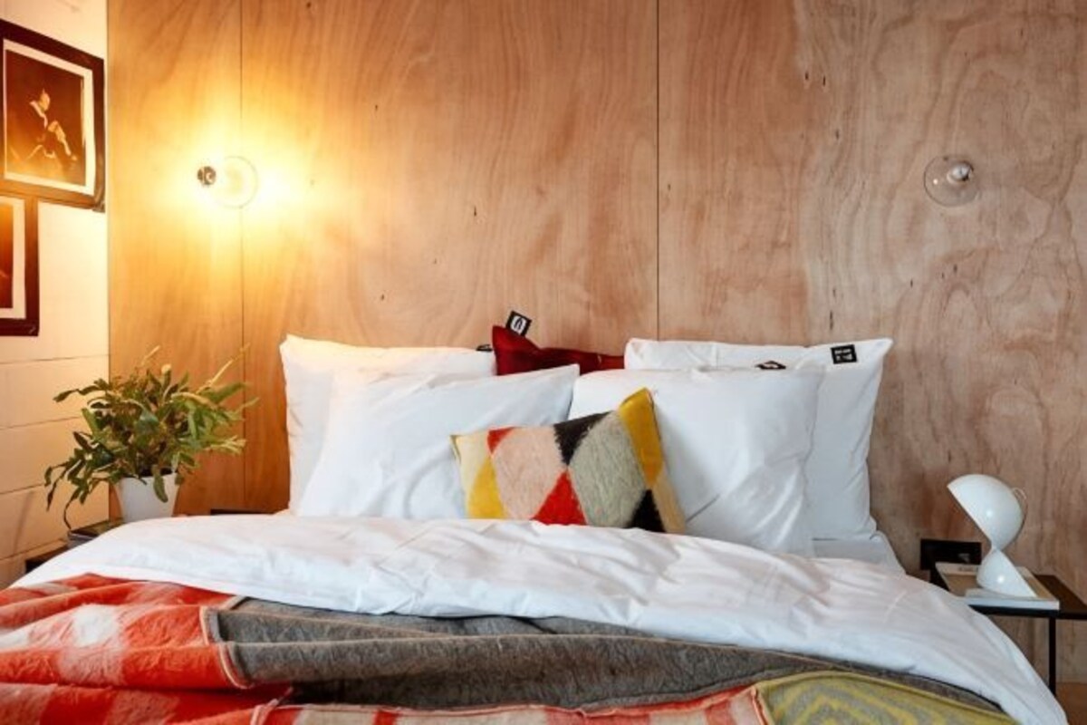 A hotel room with a wooden headboard, a bed with white bedding, a red throw blanket, a plant, and a wall-mounted light fixture.