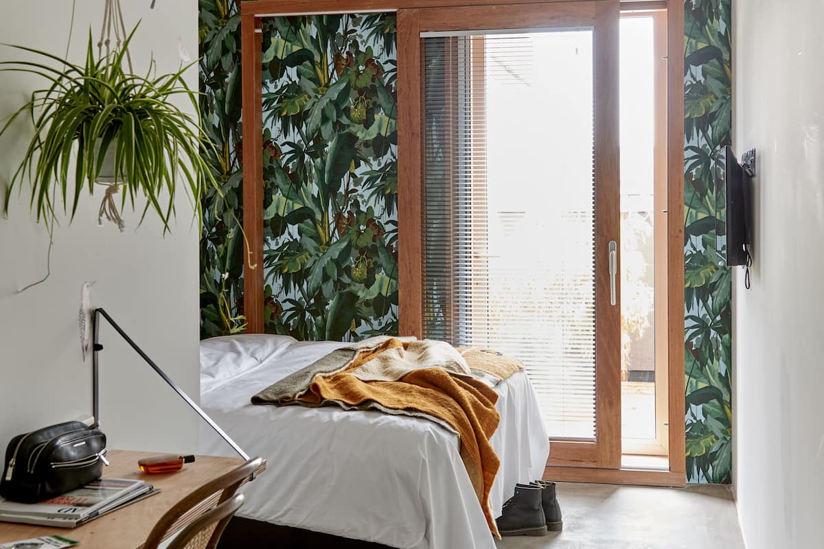 A bedroom with a bed, a desk, a chair, a plant, and a sliding door to the outside.