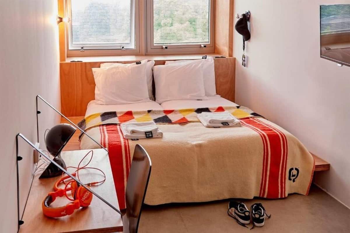 A neatly made bed with a striped blanket, a pair of headphones on a desk, and a pair of shoes on the floor.