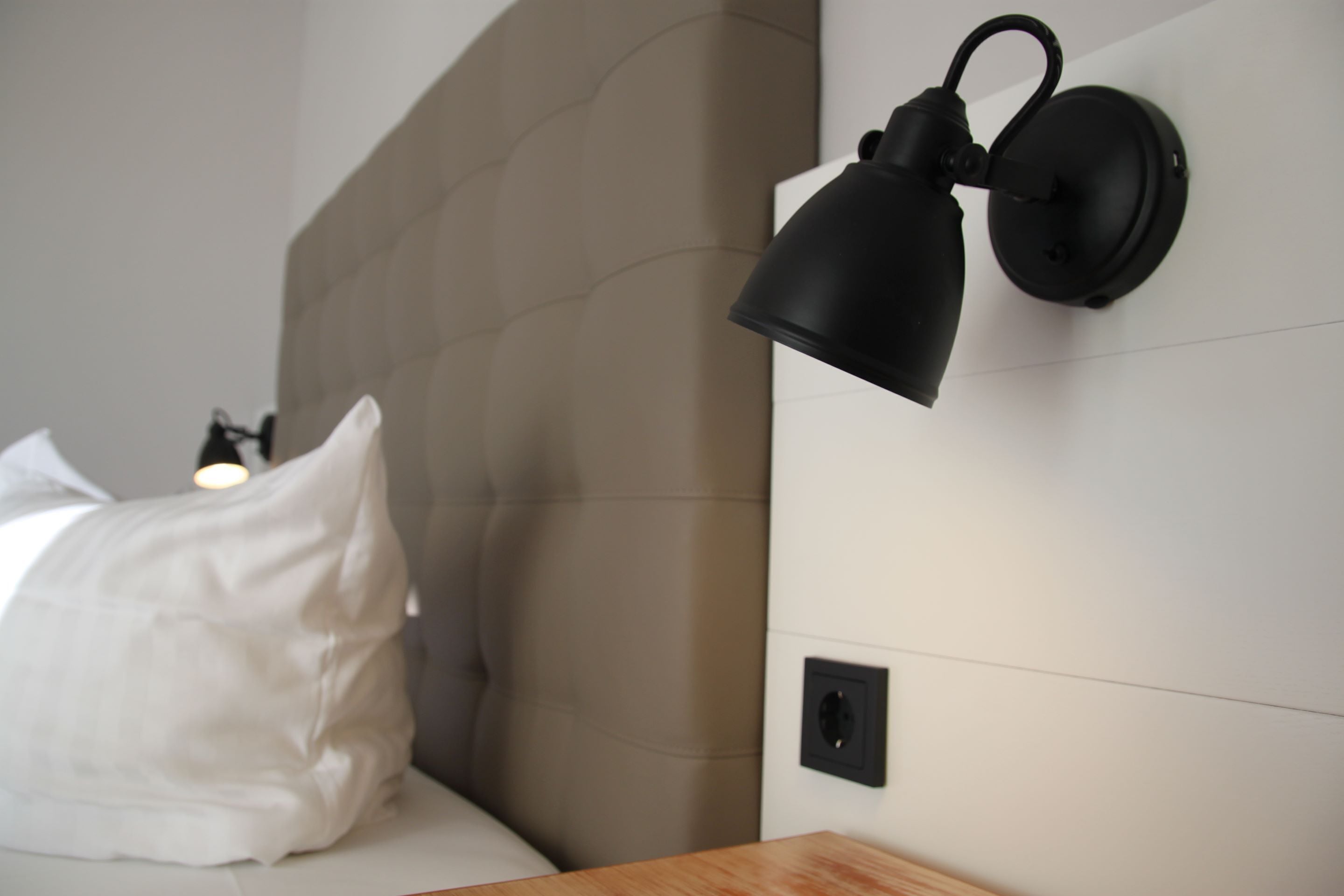 Ein ordentlich bezogenes Bett mit weißen Leinen und einem einzelnen weißen Kopfkissen, eine an der Wand befestigte schwarze Lampe und ein Nachttisch aus Holz.