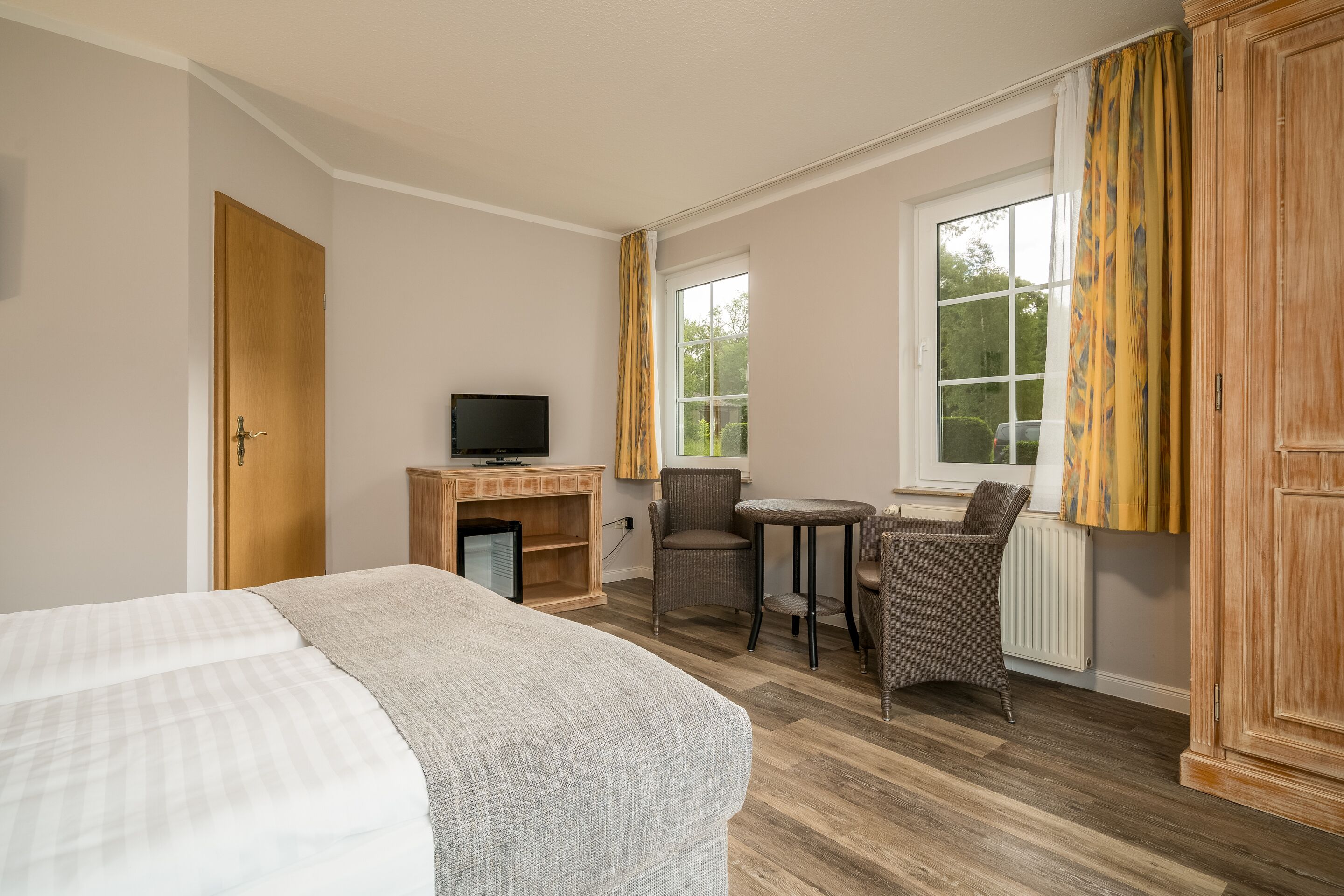 A hotel room with a bed, a television, a small table, two chairs, a window with curtains, and a wardrobe.