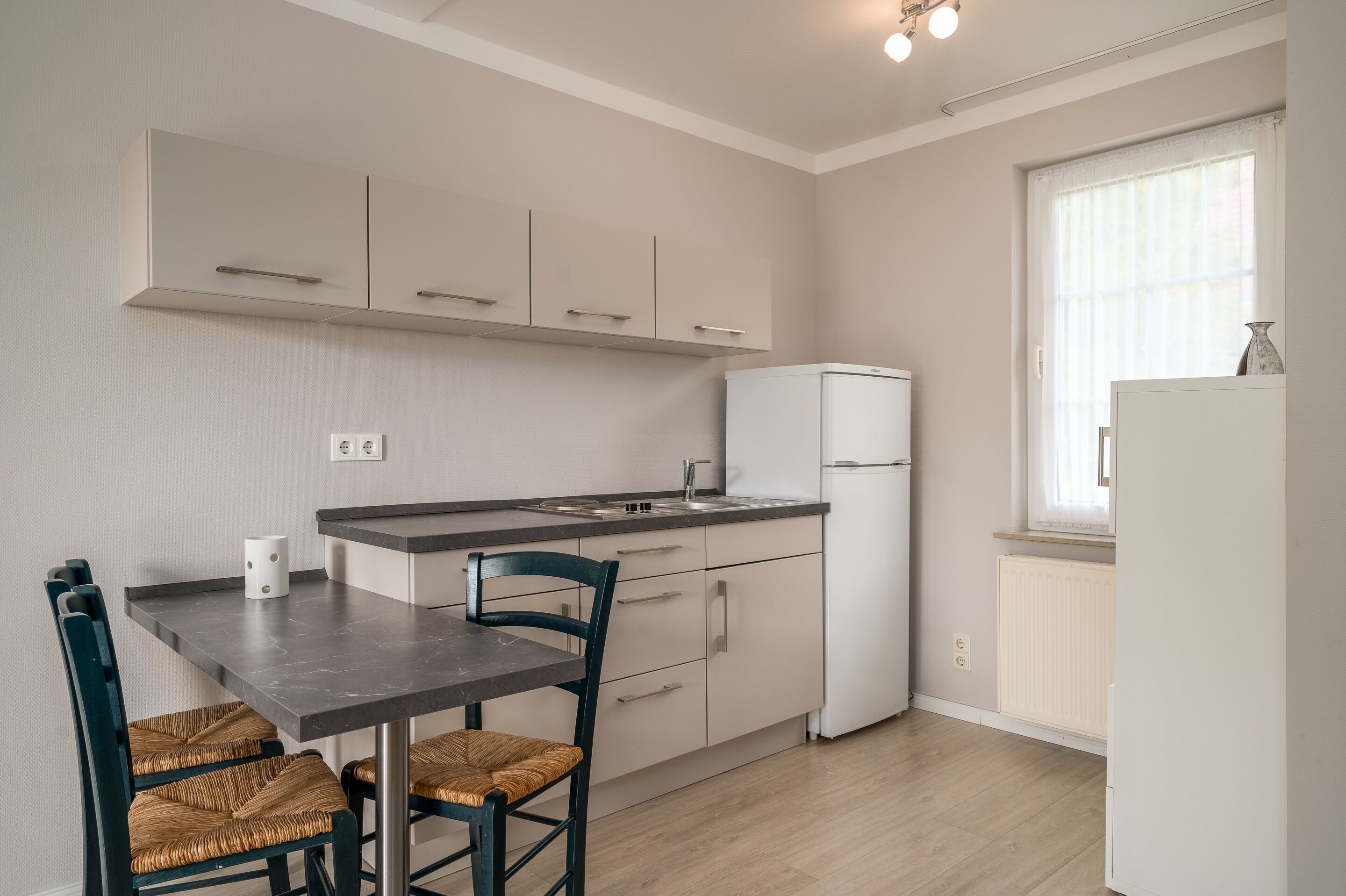 A compact kitchen with a dining table and chairs, a refrigerator, and a window with curtains.