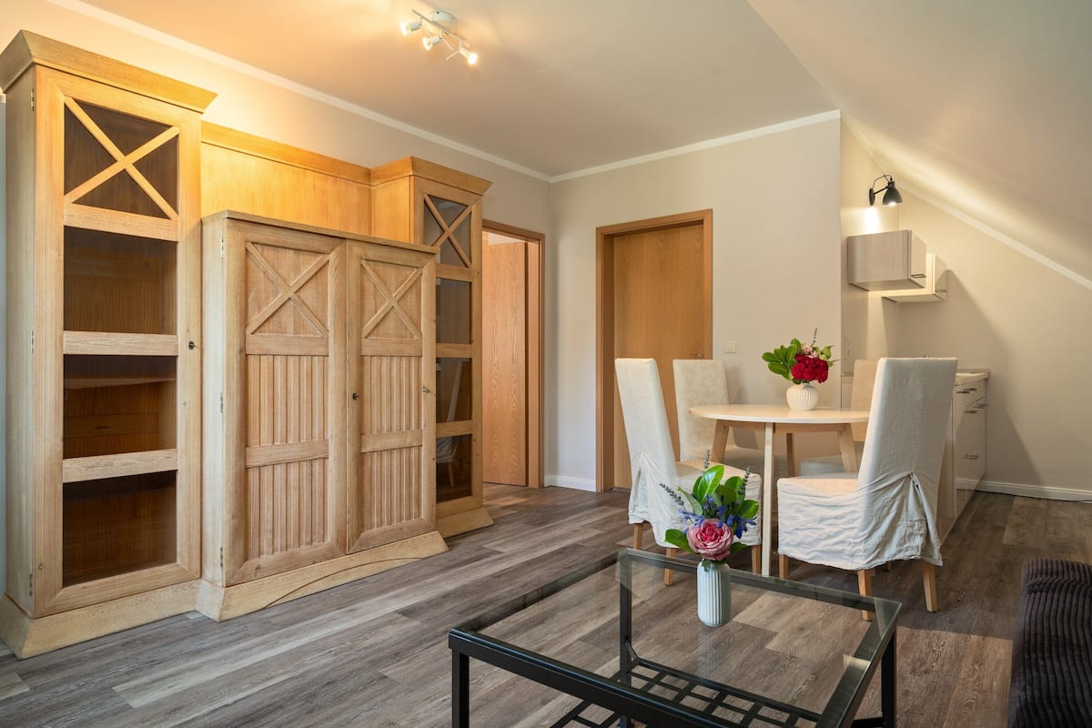 Una habitación con un armario de madera, una mesa de comedor con sillas y una mesita con un jarrón de flores.