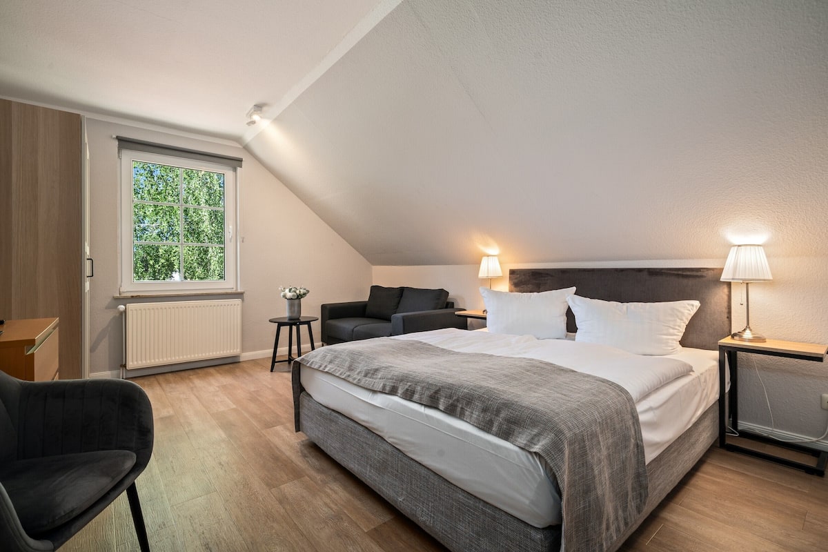 Un dormitorio moderno con una cama grande, un escritorio, una silla, un sofá y una ventana con vistas a árboles.