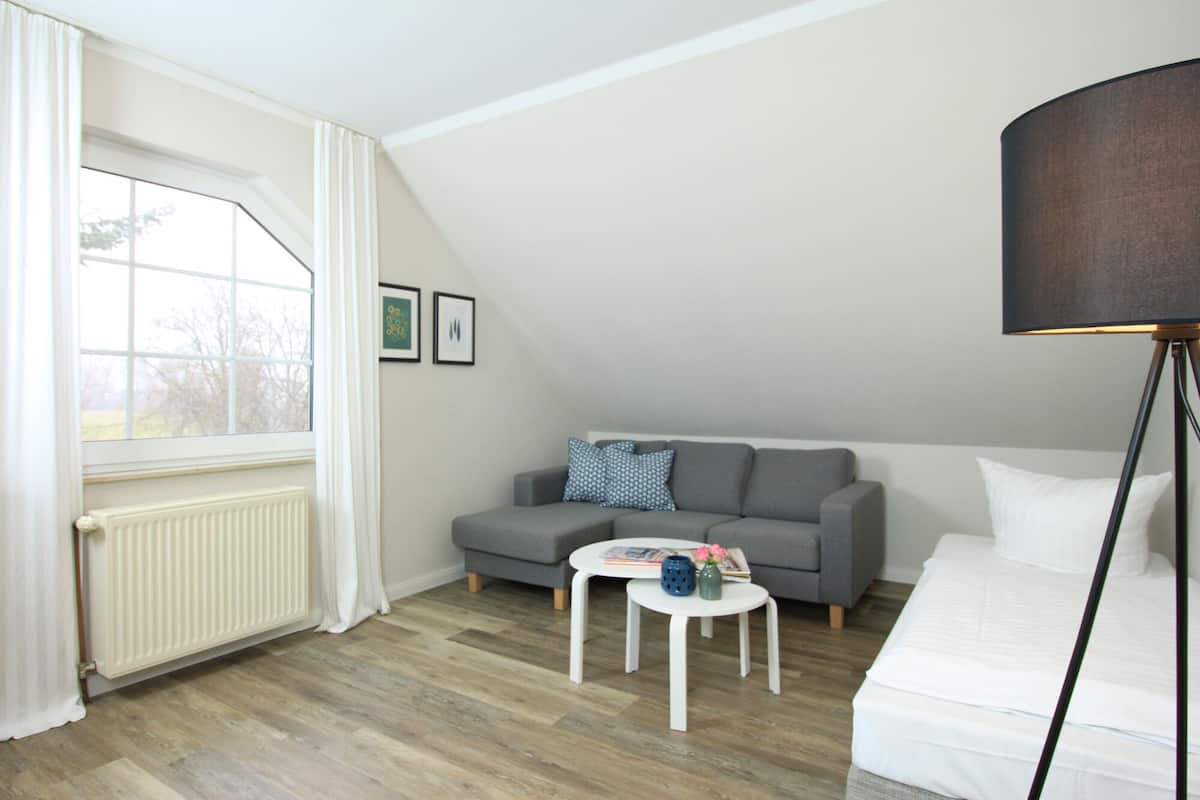 Una habitación moderna y bien iluminada con un sofá gris, una mesa de centro blanca y una lámpara de pie. Se ve un radiador a la izquierda, y hay cuadros enmarcados en la pared.