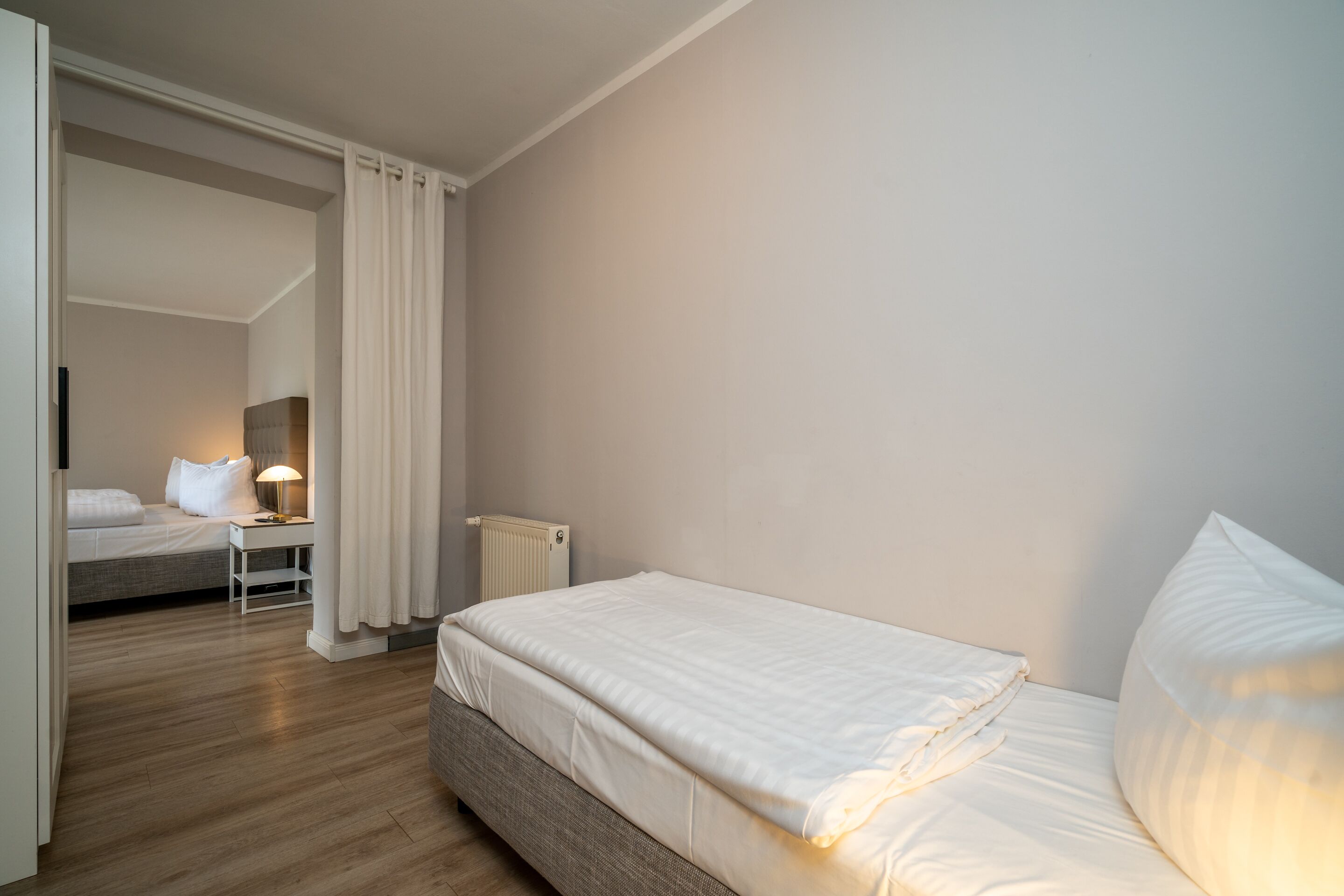 A bedroom with a single bed, a bedside table with a lamp, a radiator, and a view into another room.