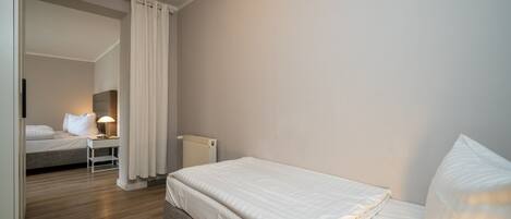 A bedroom with a single bed, a bedside table with a lamp, a radiator, and a view into another room.