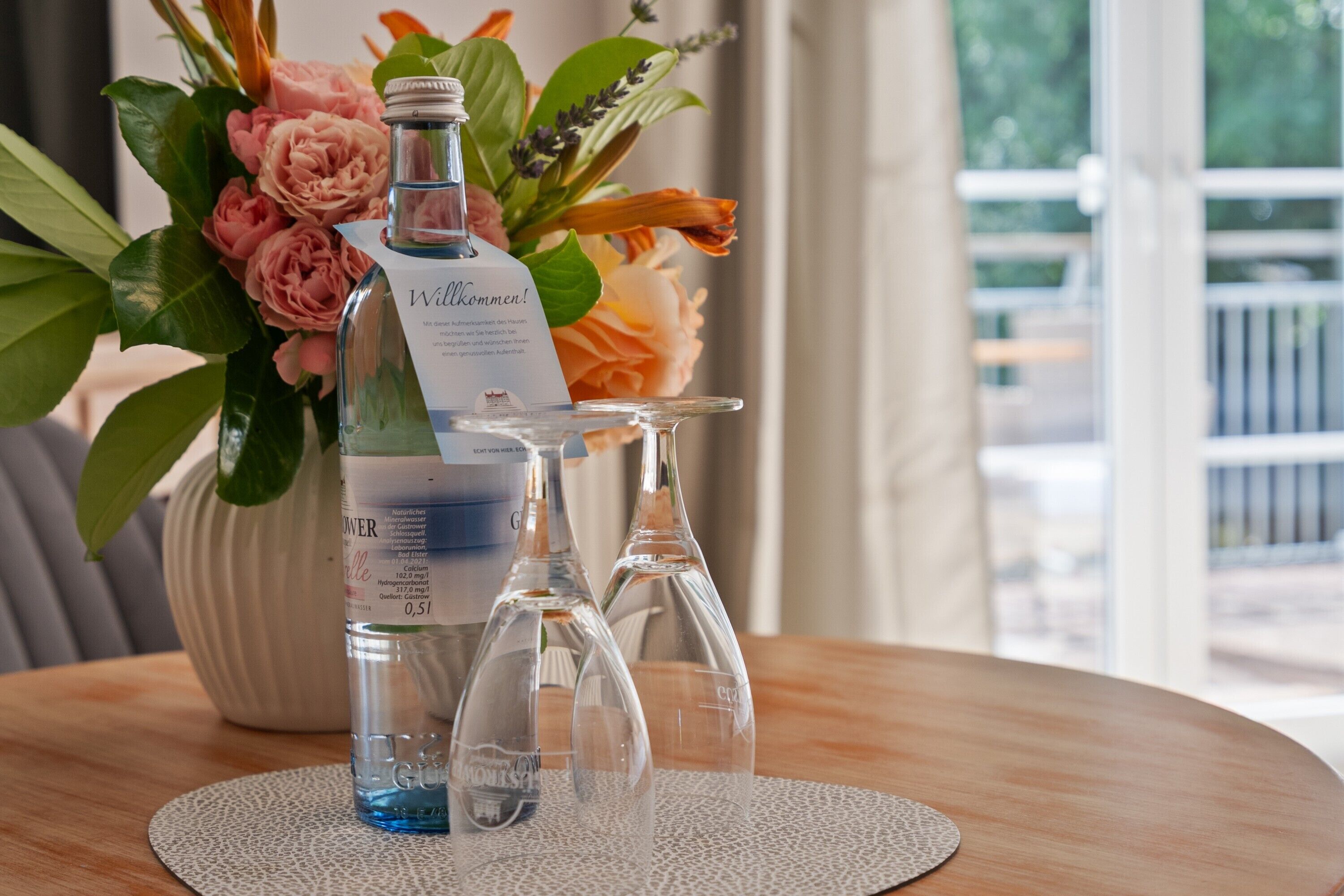 A bottle of water with a label and two empty wine glasses on a round table with a placemat.