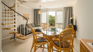 A modern living room with a glass-top dining table, wicker chairs, a grey sofa, a staircase with wooden steps, and a view of greenery outside. - Hotel Müritz - Park (Rechlin)