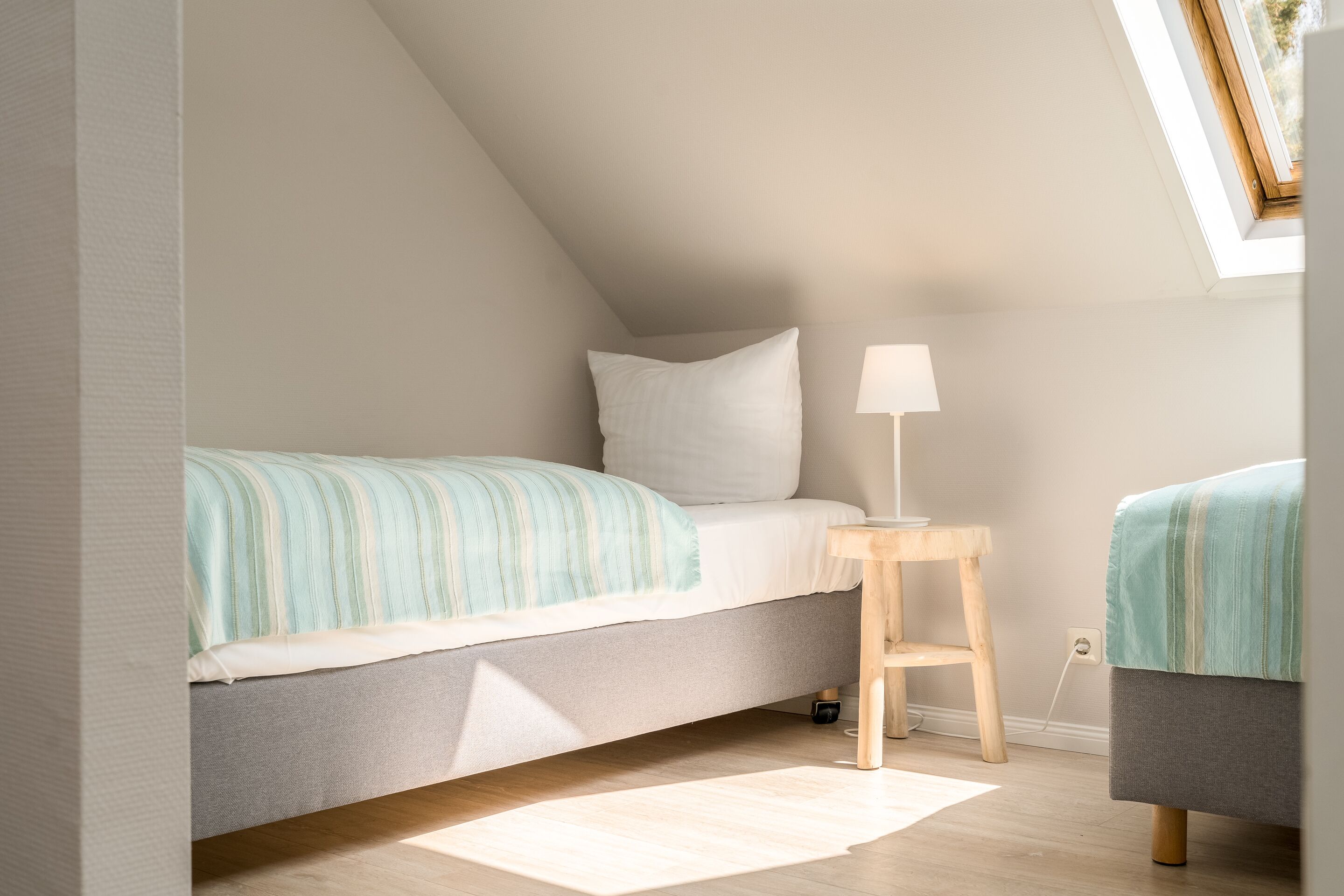 A bedroom with a sloped ceiling, a bed with a striped blanket, a bedside table with a lamp, and a small stool.