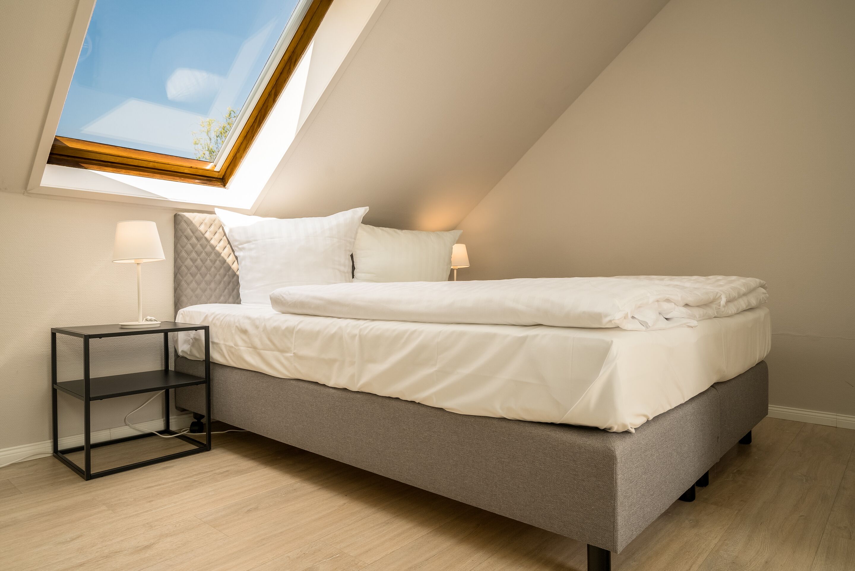 Un dormitorio moderno con techo inclinado, una cama con ropa de cama blanca, una mesita de noche con una lámpara y un tragaluz.