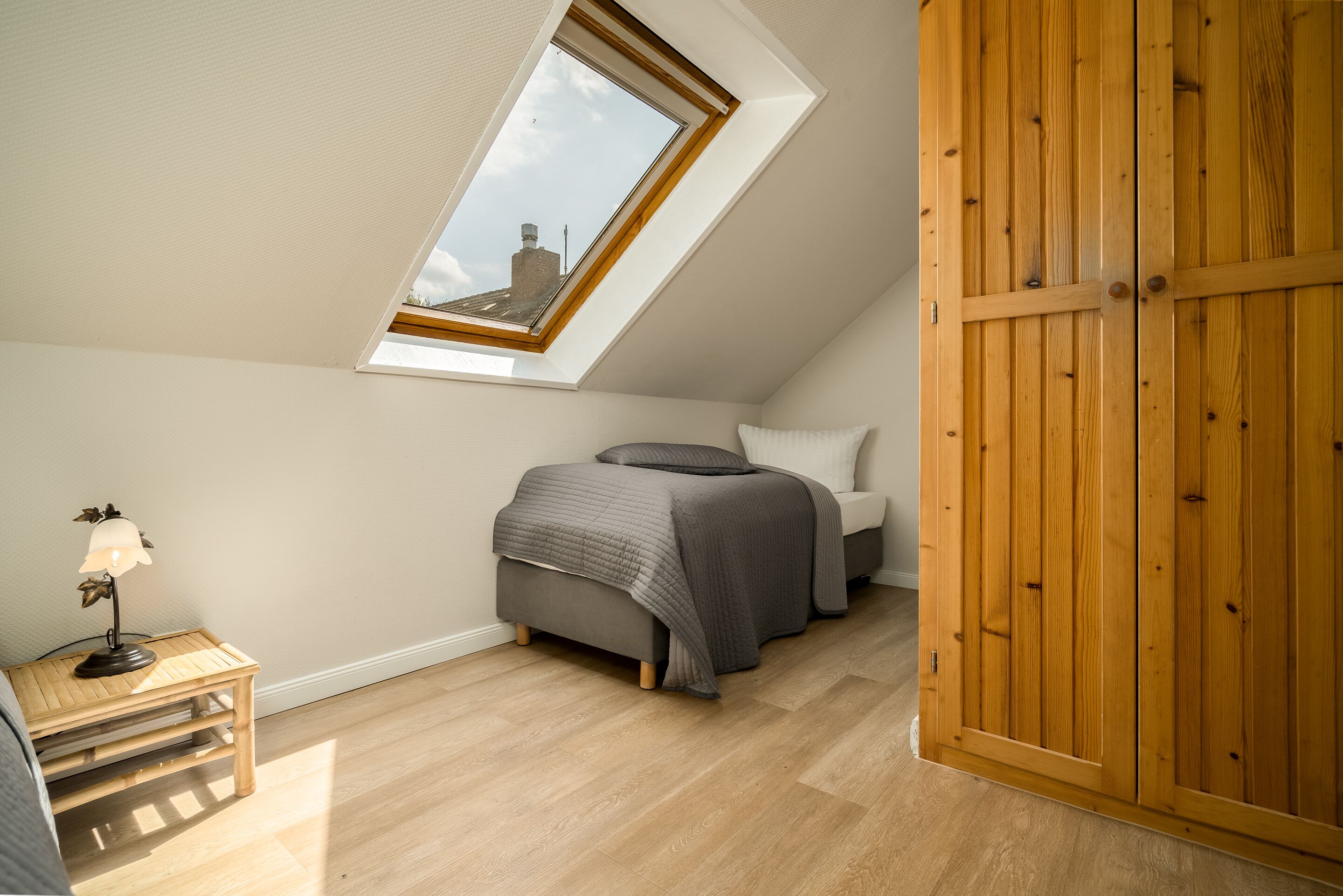 A bedroom with a sloped ceiling, a skylight, a bed with grey bedding, a bedside table with a lamp, and a wooden wardrobe.