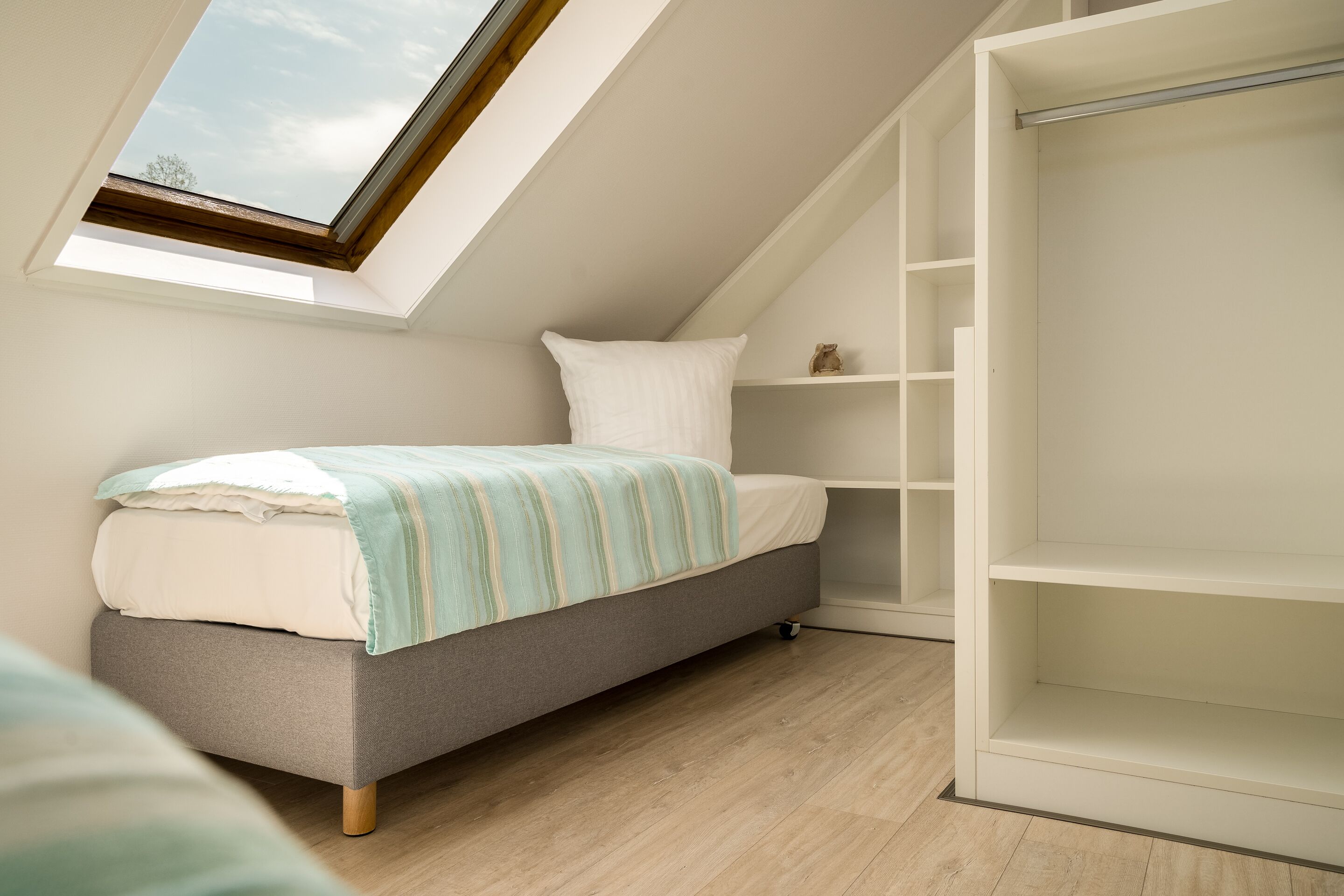 Un dormitorio con tragaluz, cama con manta a rayas, armario blanco y suelo de madera.